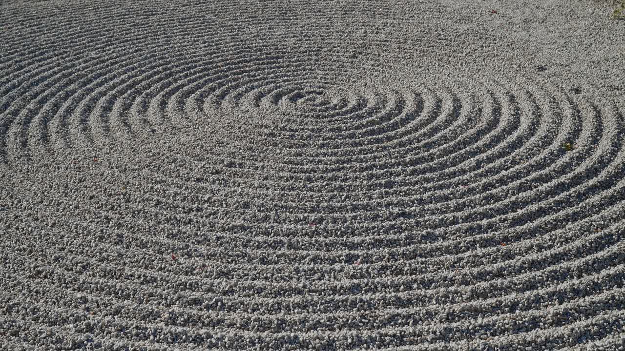 Un primo piano dettagliato di un giardino secco Karesansui. La luce del sole evidenzia gli intricati motivi circolari di ghiaia rastrellata e le increspature nella sabbia bianca, creando un'atmosfera meditativa e pacifica