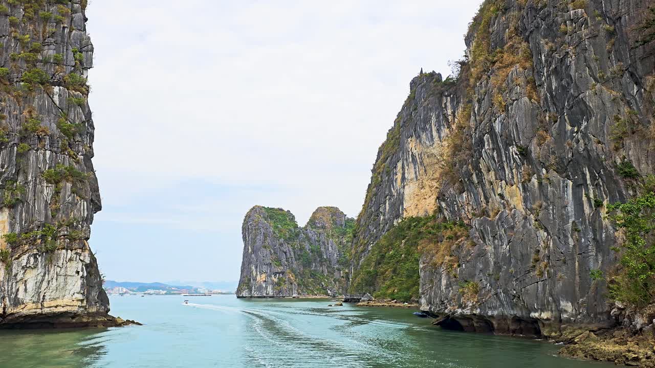 Towering limestone karsts rise dramatically from the emerald waters of Ha Long Bay, creating a majestic natural gateway where tourist boats navigate through the tranquil channel between the cliffs