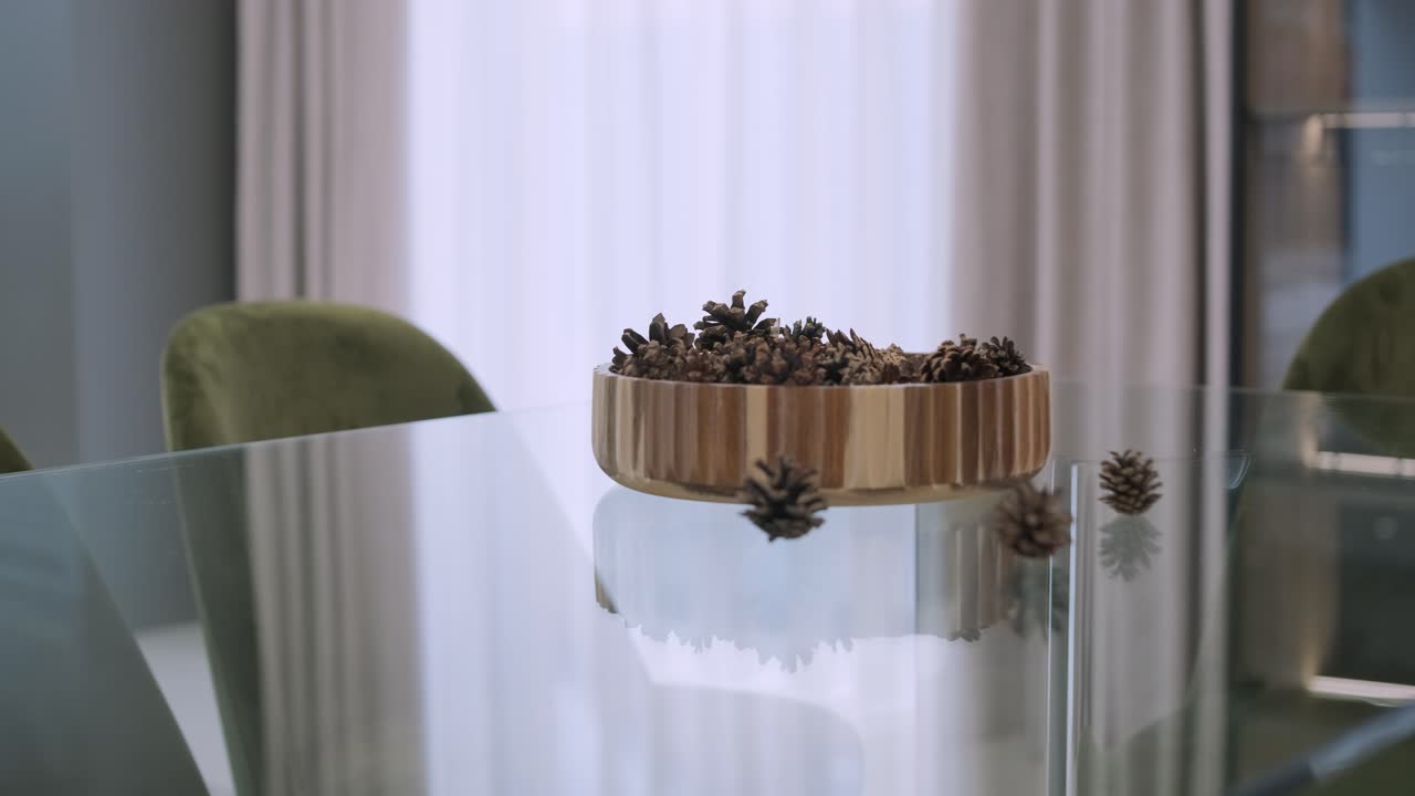 On a sleek glass table, a wooden bowl filled with pine cones makes a simple yet elegant natural centerpiece, reflecting the minimalist charm of the space