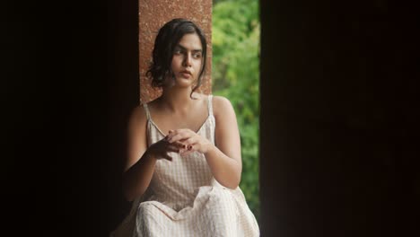 Mujer-Joven-Con-Un-Vestido-De-Verano-Sentada-Contemplativamente-Junto-A-Una-Ventana-Rústica,-Luz-Natural-Suave