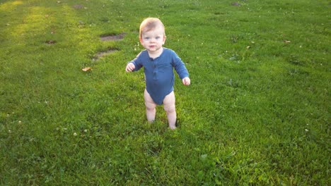 happy toddler boy stands up alone in grass field, almost loses balance, and smiles