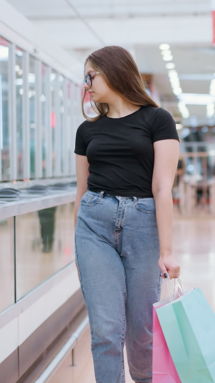 mujer de compras en la parte superior negra camina más allá del vidrio de exhibición con otro comprador en el fondo, ella sostiene bolsas de compras, mirando hacia adelante mientras se mueve a través de un ambiente de centro comercial brillante y moderno