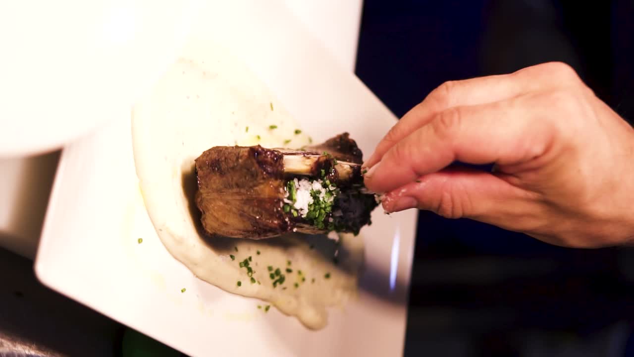 toma vertical en cámara lenta de costillas sabrosas adornadas con hierbas y echadas en una salsa blanca mientras un chef agrega sal a la carne en un restaurante fino