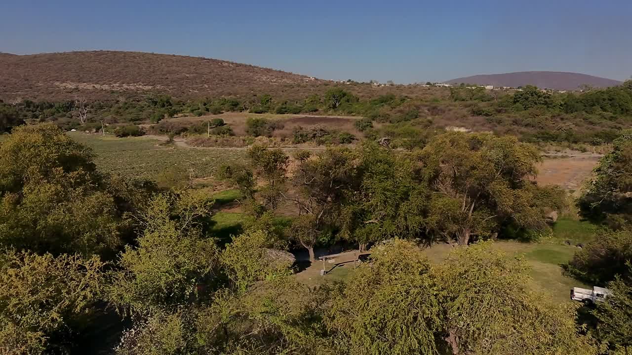 Backward drone flight over green tree canopy near a riverbank with a visible camping area below, surrounded by hilly terrain under clear skies.