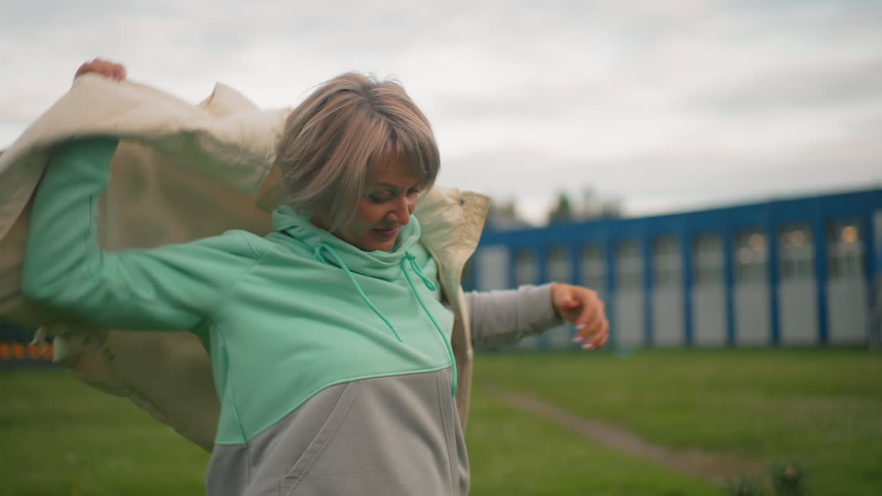 Graceful middle aged woman with soft smile wears sleeveless coat over mint green hoodie while enjoying fresh outdoor air in open grassy field beside modern building under cloudy sky