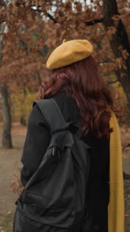vista trasera de una mujer con muffler amarillo y boina caminando por el bosque, sosteniendo la correa de la bolsa y mirando hacia el lado, el cabello balanceándose en la brisa de otoño, la figura borrosa en el fondo