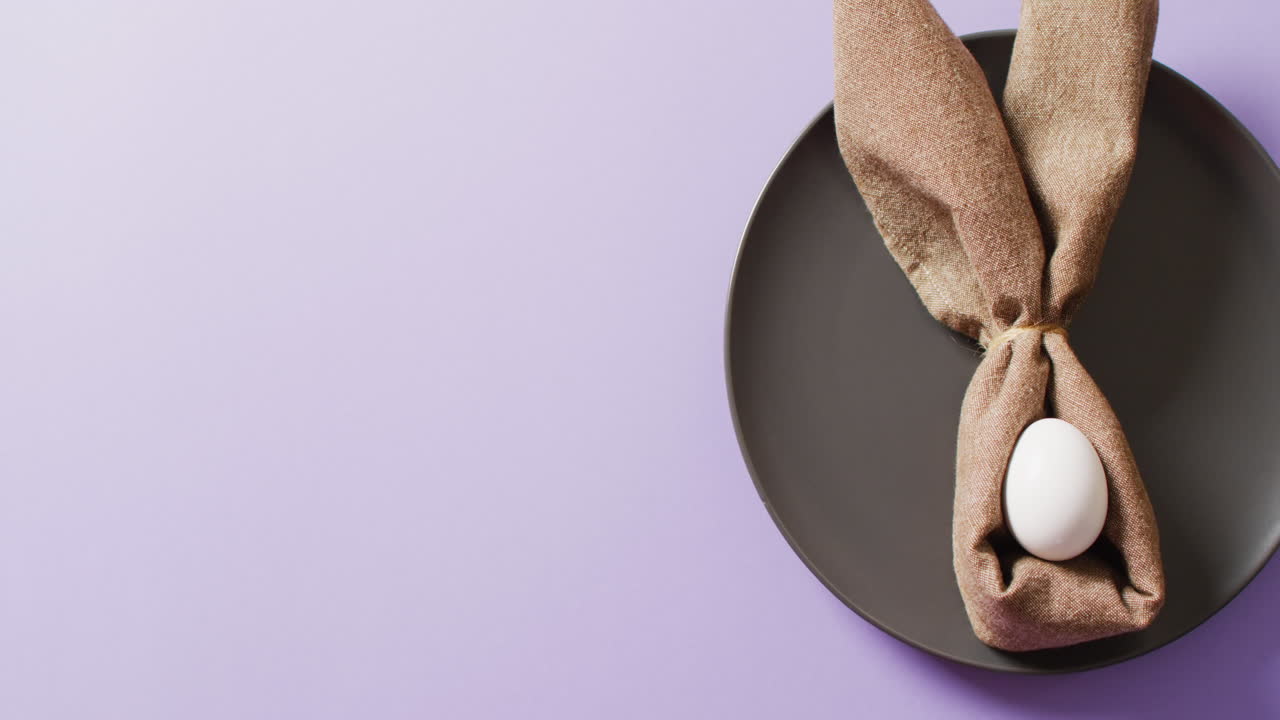 Egg and ribbon in shape of rabbit on plate on purple background with copy space