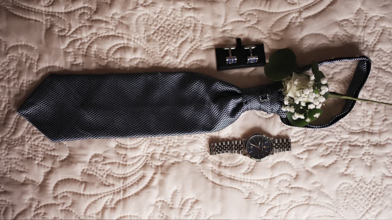 groom's tie watch cufflinks and boutonniere arranged on beige textured bedspread