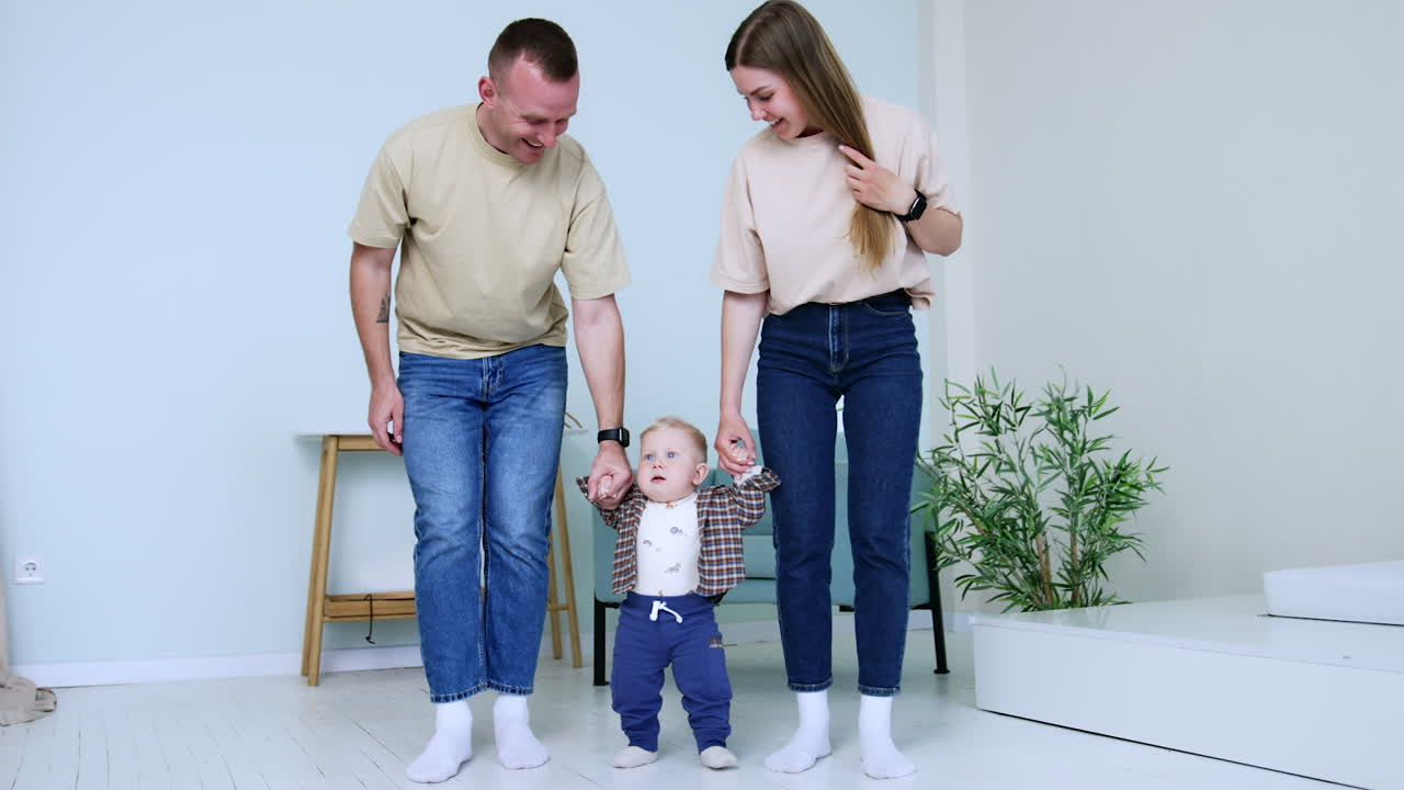 Parents hold the hands of their little infant baby stepping by the floor. Mom and dad teaching their son to walk.