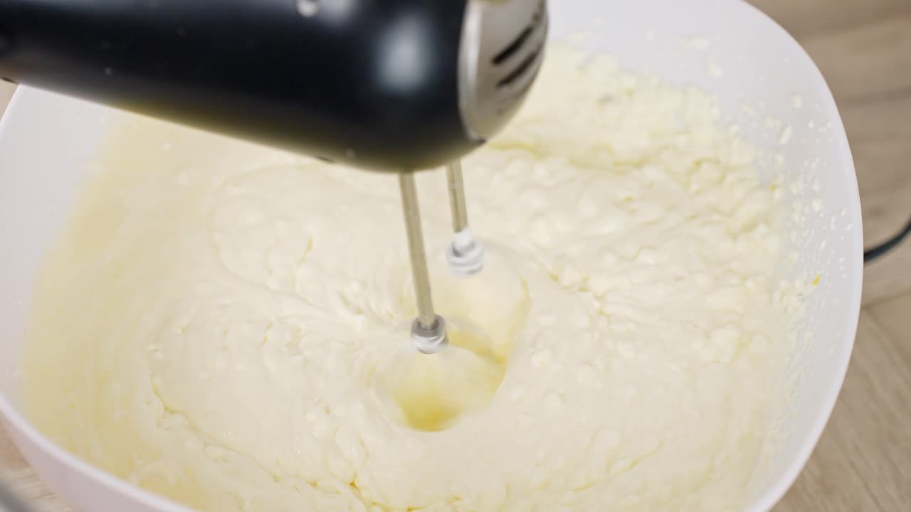 Close-up shows mixer beating tiramisu cream made with mascarpone, egg yolk and sugar as it thickens into smooth consistency in white bowl over wooden surface, real time, static kitchen prep shot