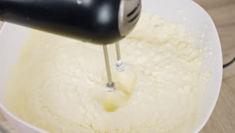 close-up shows mixer beating tiramisu cream made with mascarpone, egg yolk and sugar as it thickens into smooth consistency in white bowl over wooden surface, real time, static kitchen prep shot