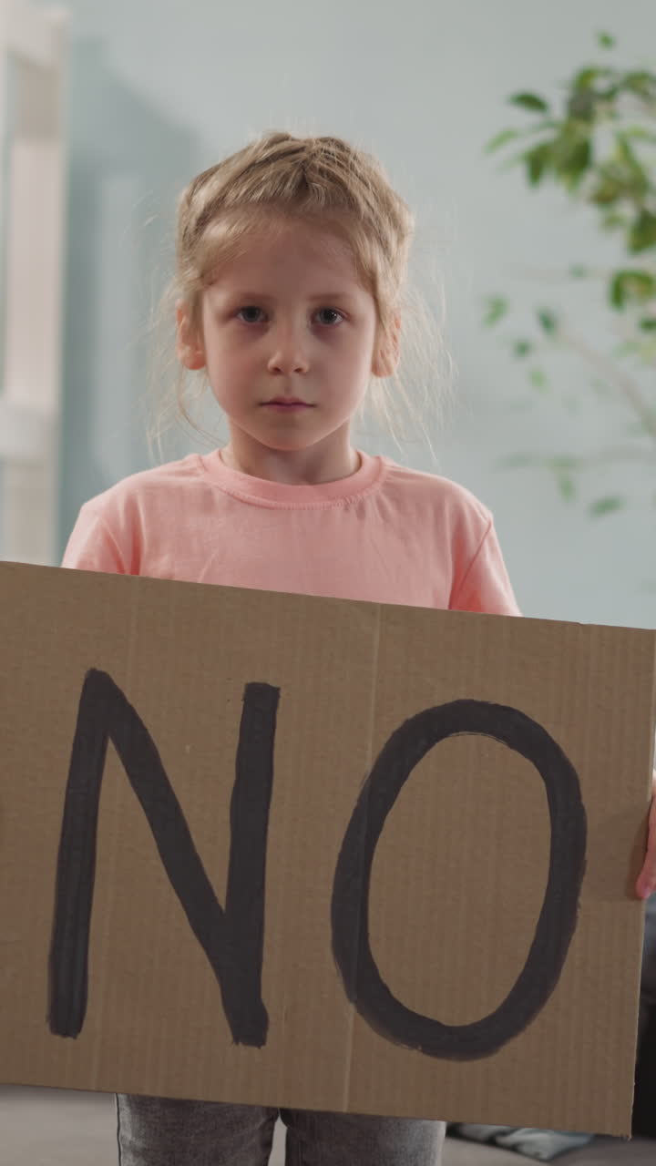 tired sad resentful little girl stands and holds poster with inscription NO in her hands, child protests in room near window