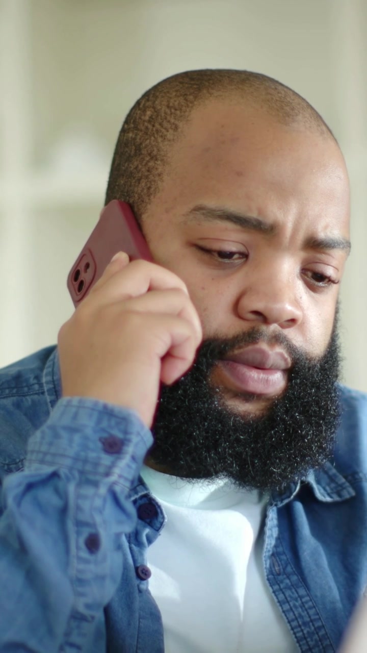 Vertical video: Taking call, man in denim at home holding maroon phone and checking laptop for work