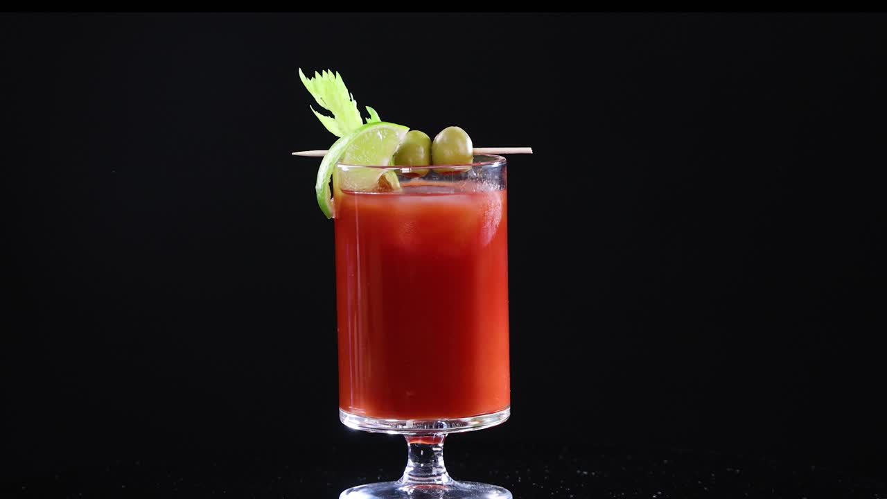 A hand garnishes a Bloody Mary cocktail with olives and celery against a black background, highlighting the drink's vibrant colors