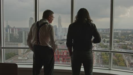 wide view of an older businessman points towards the city skyline of rotterdam, the netherlands while talking to an employee