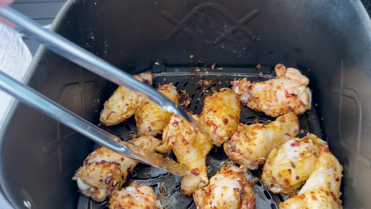 Hand turns cooked chicken wings with metal tongs inside an air fryer under bright lighting