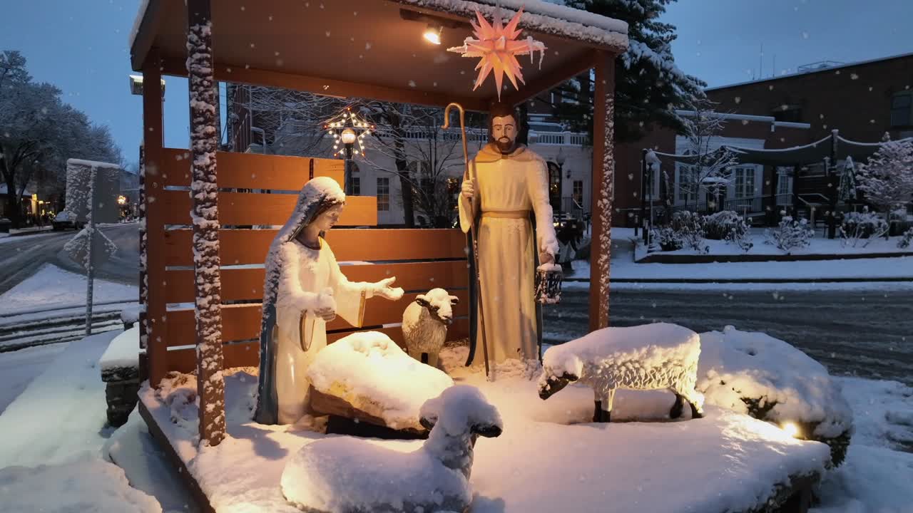 Scena di una città americana innevata con tradizionale presepe natalizio al crepuscolo, figure illuminate delicatamente e circondate da neve che cade. Atmosfera pacifica e festosa di vacanza invernale. Ripresa aerea ascendente