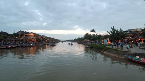 lanterns by a river with many people walking by and shops decorated with lights