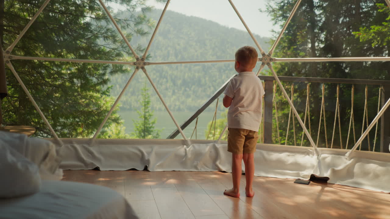 Child stands alone under glass dome at glamping. Thoughtful little boy looks out of panoramic window on terrace. Kid observes calm nature scene