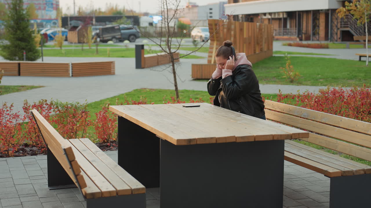 Young girl seated at wooden table in leather jacket preparing to wear headphone outdoors as red shrubs sway gently in wind with parked vehicles and modern buildings in background