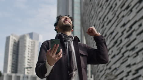 Hispanic-ethnicity-man-cheers-up-happily-receiving-good-news-while-on-a-video-call-outdoors-on-a-sunny-afternoon-in-downtown