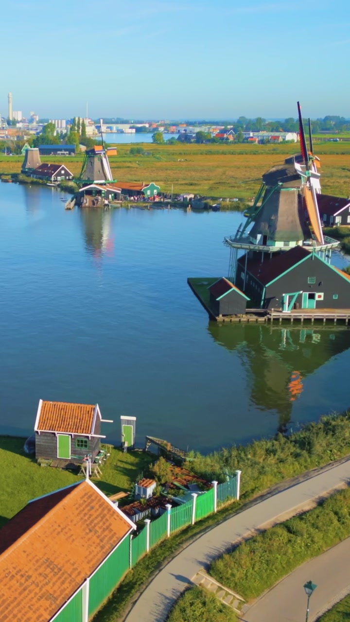 Aerial view of iconic windmills and traditional houses at Zaanse Schans, Zaandam, Netherlands. The vibrant green structures stand by tranquil canals under a clear blue sky, showcasing Dutch heritage.
