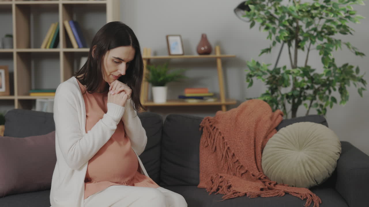 Pregnant woman sitting on sofa, appearing pensive and thoughtful.