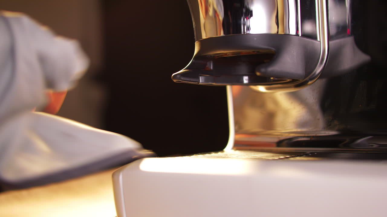 Primer plano de una mano limpiando el cabezal de preparación de metal y la zona del portafiltro de una brillante máquina de espresso. La iluminación suave y los tonos cálidos enfatizan la limpieza y el cuidado en el entorno de preparación de un barista