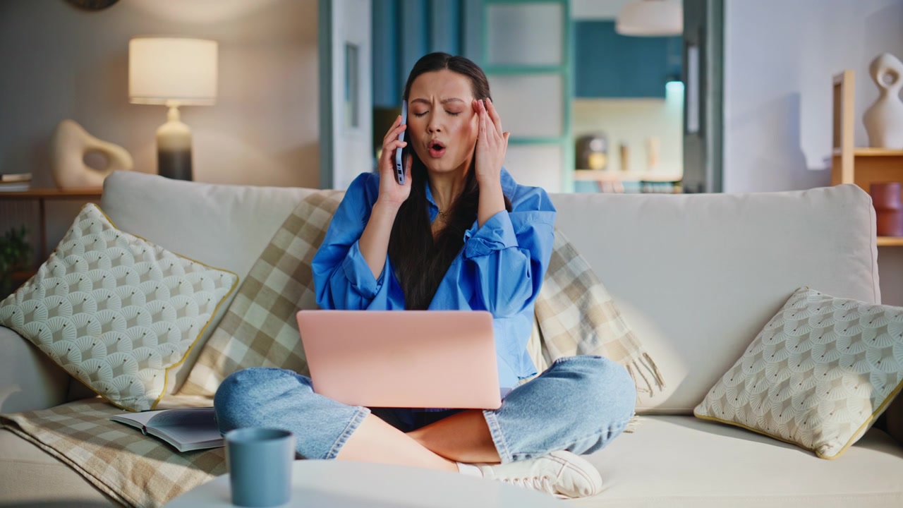 Frustrated asian woman talking phone working laptop sofa. Stress headache home