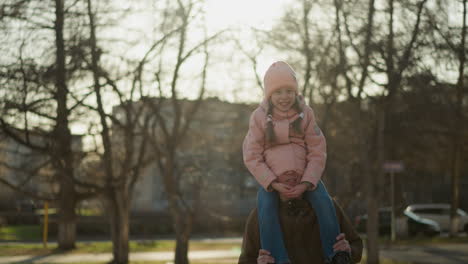 a little girl in a pink cap and jacket sits joyfully on her dad's neck, with half of his head snugly tucked in her jacket covering his eyes as they smile joyfully