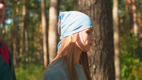 close-up of sisters walking through sunlit forest, one wearing red backpack with cloth draped over it, other with blue headscarf, sunlight filters through trees, casting a soft glow on their faces