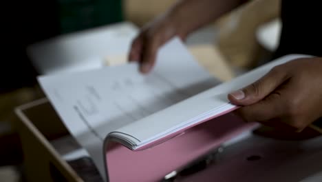 person flipping through white documents organized in a pink binder, likely involved in organizing or searching for important information or files