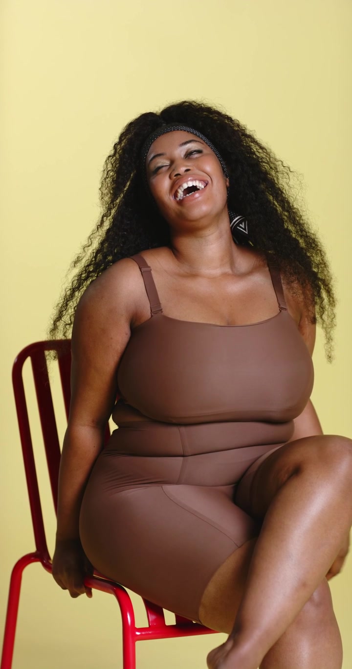 Video verticale: Donna afroamericana che, seguendo un'indicazione, accavalla le gambe su una sedia rossa in studio, ridendo.