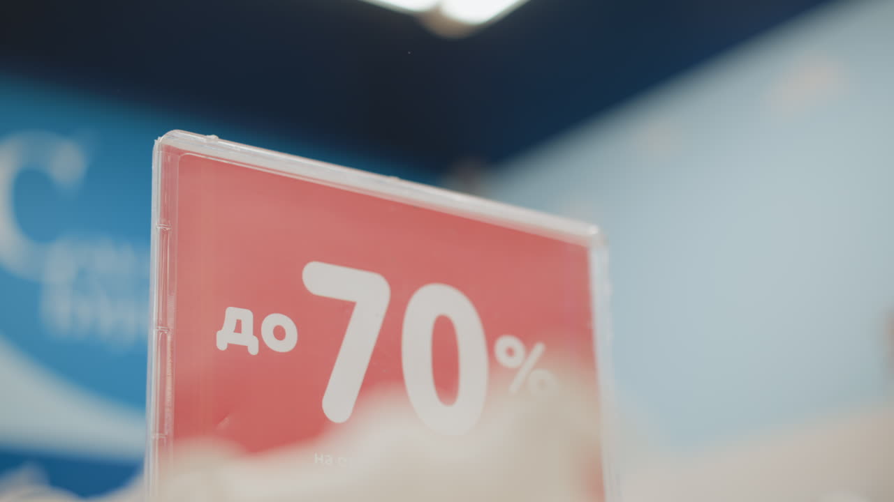 Price sign displaying up to seventy percent discount in retail clothing store, red board with white typography, blurred jackets and racks in background, promotional display for sale