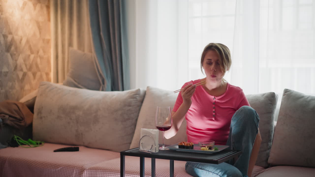 Young girl seated comfortably on sofa in cozy home setting enjoying dinner with chopsticks and wine as warm golden light shines through curtains highlighting relaxed casual evening atmosphere