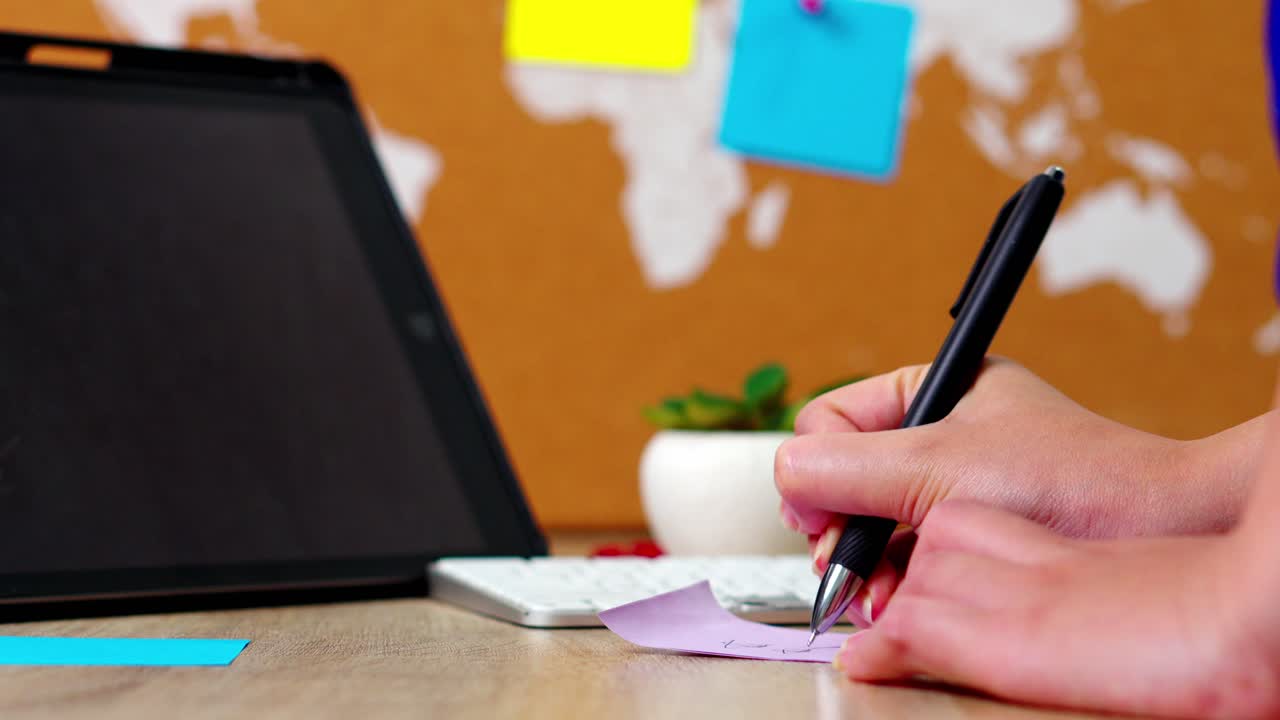 Close-up of hands writing to-do list on purple sticky note at wooden desk with laptop and world map