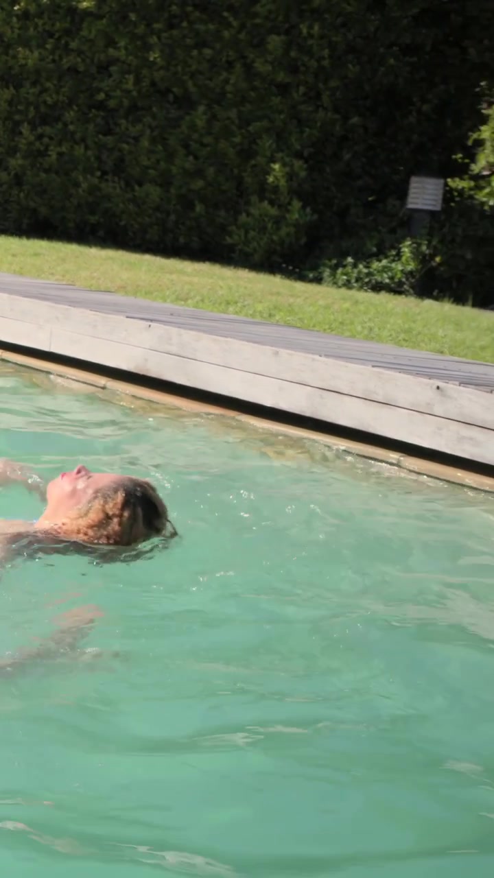 Vertical video: Pushing off pink ring, blond woman heading to deck at backyard pool in swimwear