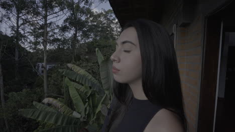 beautiful, young, happy woman in her 20's wearing black top with jeans stares into the distance from balcony with plants in background