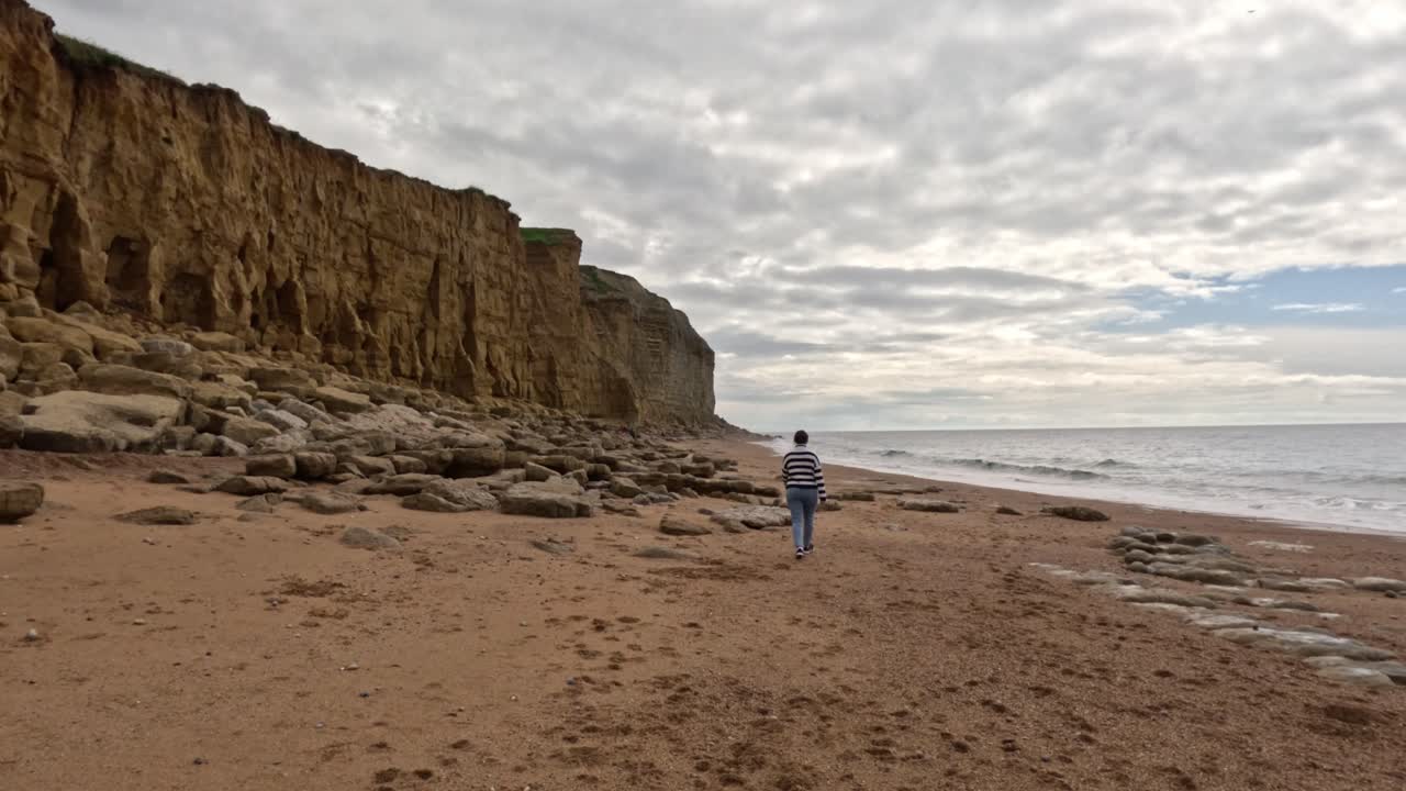 The camera moves sideways to the right as the female walks away along the beach wide the sea on the right and the cliffs left. A women walking along a Jurassic Coast beach in Dorset