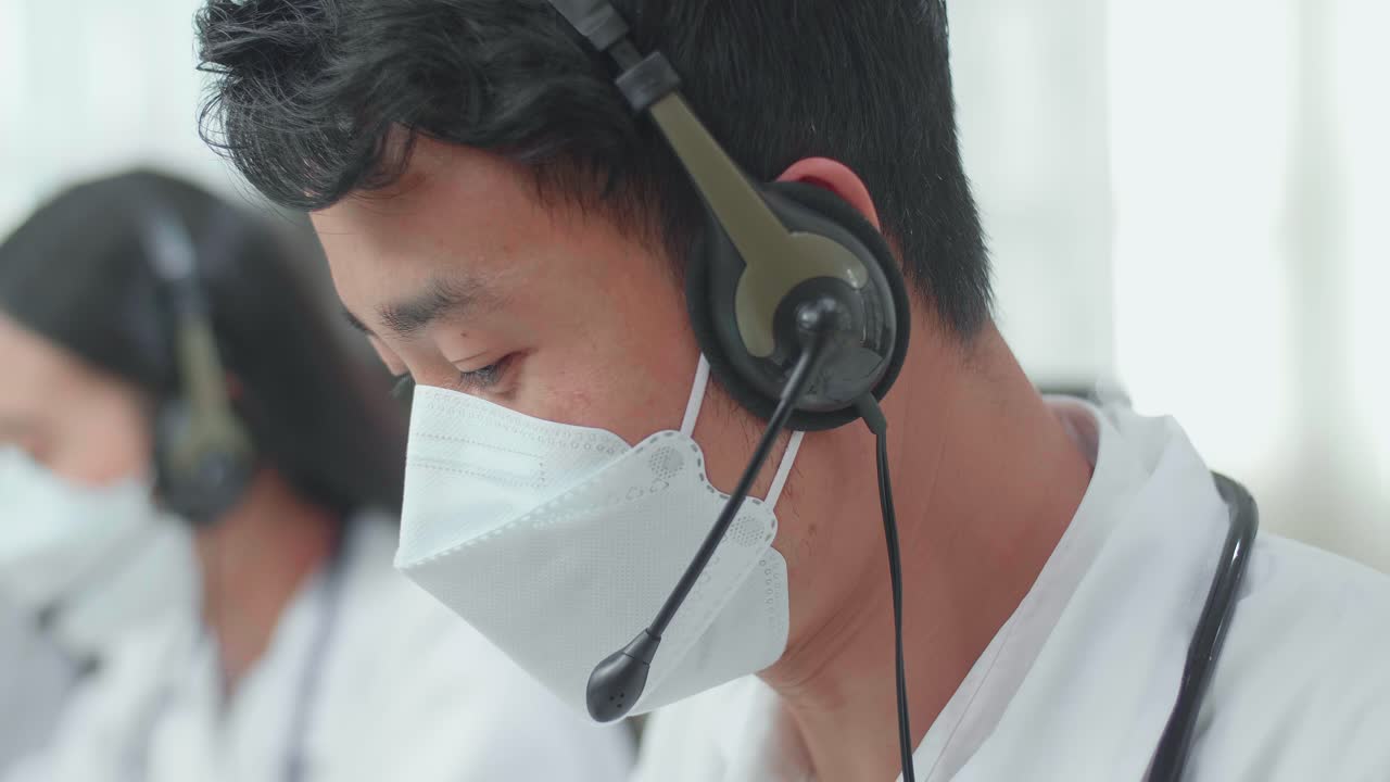 Close Up Of An Asian Man Doctor Wearing Headsets And Masks Working As Call Center Agent And Speaking To Customer On The Call During Woking With Colleagues At The Office