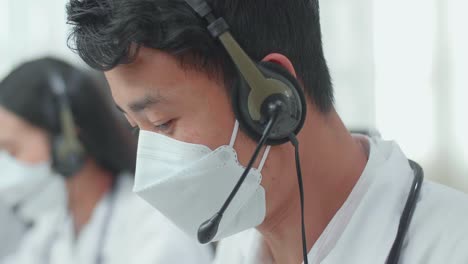 close up of an asian man doctor wearing headsets and masks working as call center agent and speaking to customer on the call during woking with colleagues at the office