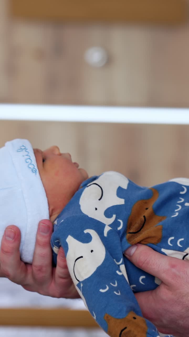 Adorabile neonato in pagliaccetto blu e berretto bianco. Il padre tiene in braccio il bambino di fronte a sé. Video verticale