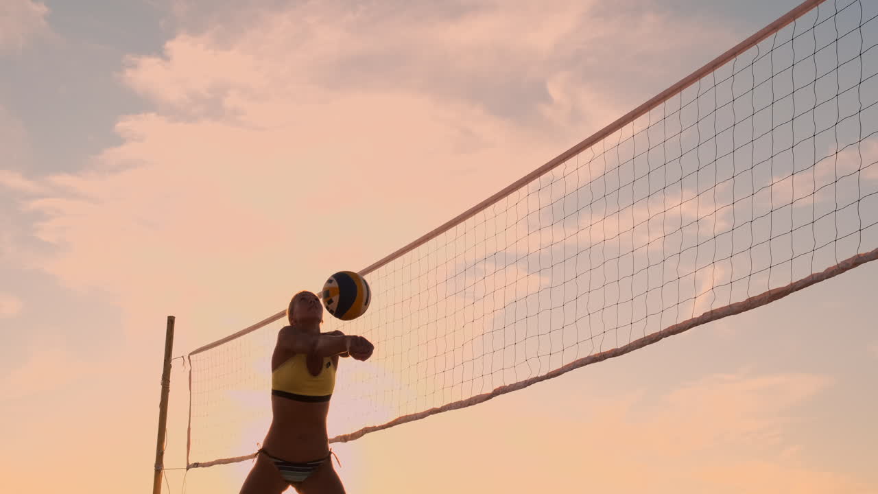 해변 볼리볼을 하는 운동선수 소녀는 공중으로 뛰어내리고 아름다운 여름 저녁에 네트 위에 공을 때립니다.