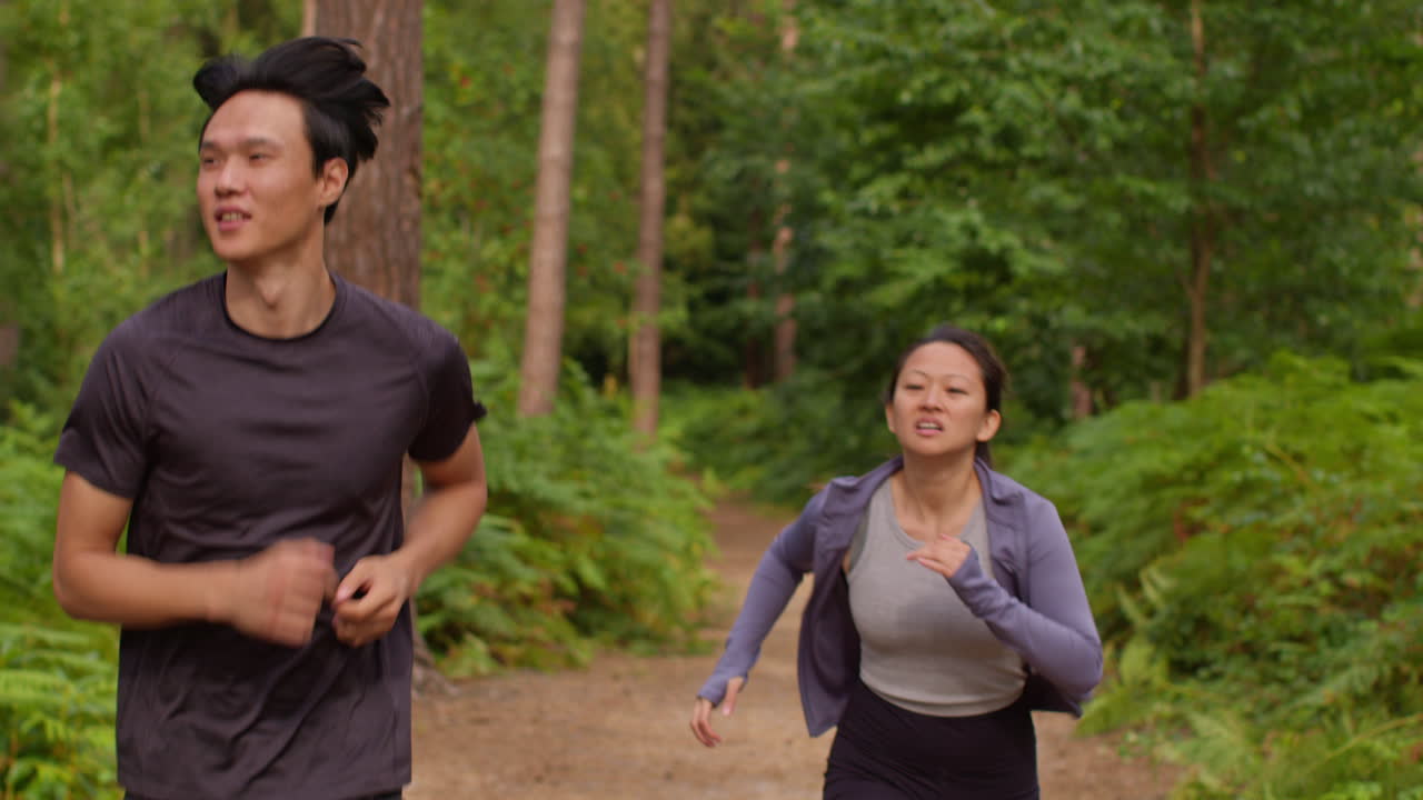 성인 부부가 운동을 하고 야외에서 운동을 하고 숲을 가로질러 달리는 남자와 함께 운동복을 입은 피곤한 여자를 격려합니다.