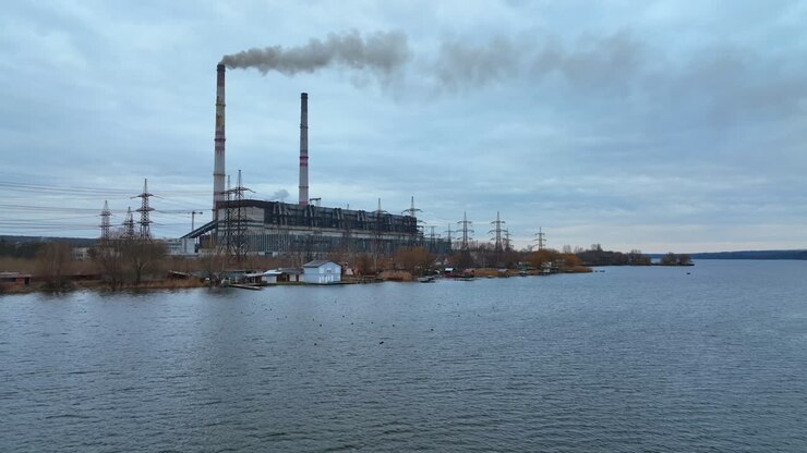 Industrial plant with two big flues rising in the air on the river bank. Dark smoke coming from the chimneys. Ducks floating on the river at the foreground.