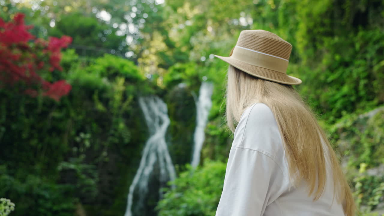 Woman in Hat Admiring Waterfall in Lush Green Landscape