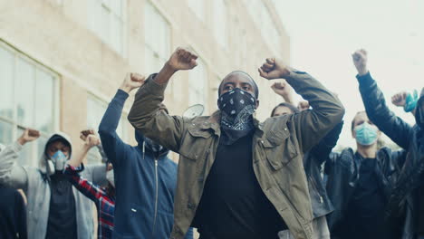 afroamerikanischer mann mit schal im gesicht und schreiend mit erhobenen armen bei einem protest mit multiethnischer gruppe von menschen auf der straße