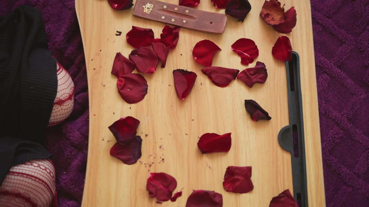 Rose petals scattered on tray, mystic reader arranges petals with wooden wand over wooden board, tarot cards visible at edge, purple cloth backdrop, closeup hands, sensual atmospheric ritual for love