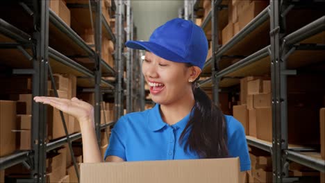 close up de mensajera asiática en uniforme azul sonriendo y señalando al lado mientras entrega una caja en el almacén