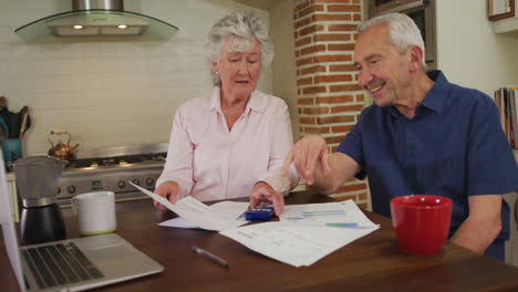 senior caucasian couple sitting in kitchen checking paperwork and using laptop computer in slow moti